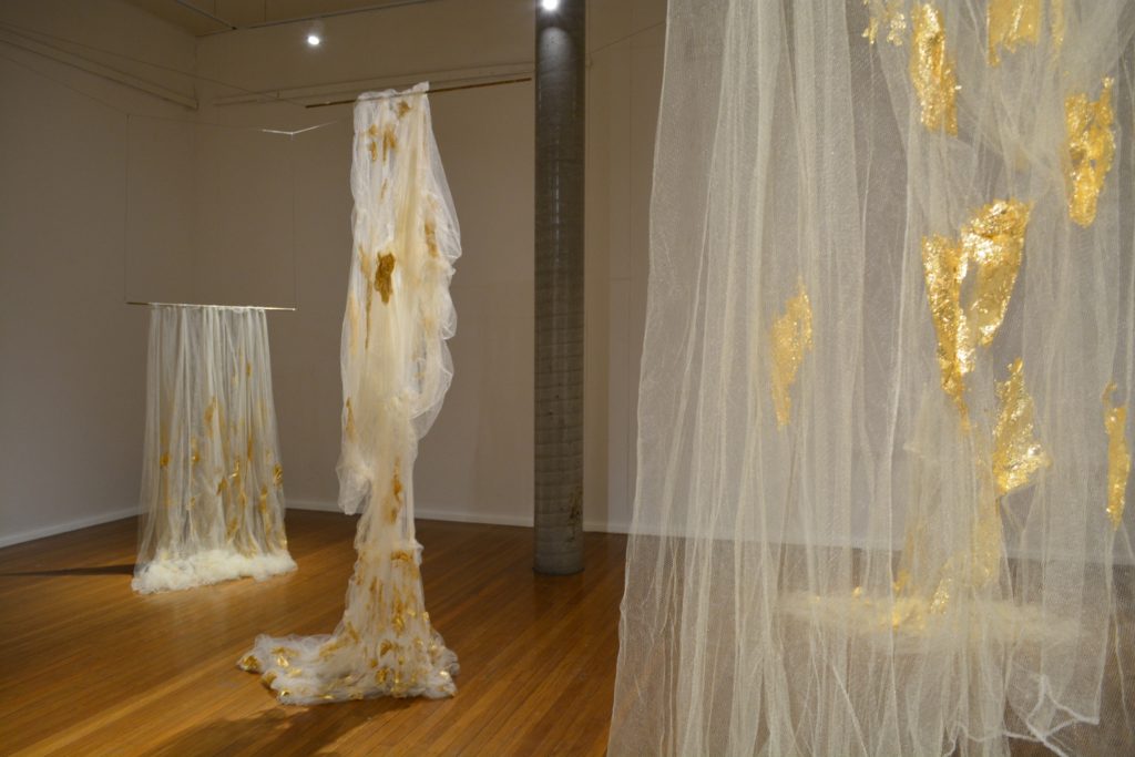 installation shot of Linda Sok's Soft Monument; 3 draped mosquito nets with gold leaf applied to them are shown