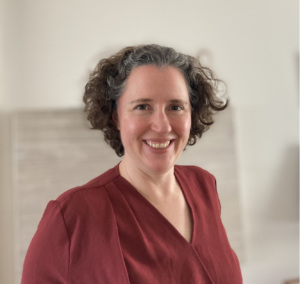 Image of Jessica May. Jessica has short, curly brown hair, and a smiling face. She wears a v neck, maroon top, and is standing in front of blank background